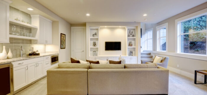 Modern basement features a gray sectional facing a white built-in tv cabinet and wet bar mounted to a wall with light colored couch and large windows where natural light comes in